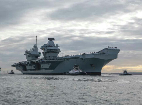 Aircraft carrier with tugboats on a cloudy sea.