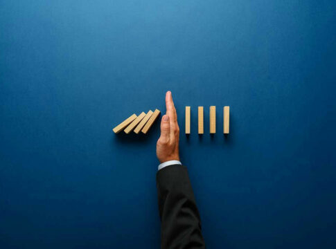Hand stopping dominoes from falling on blue background.
