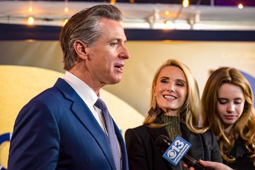 A man in a suit speaking at an event with two women nearby, one smiling and the other holding a microphone