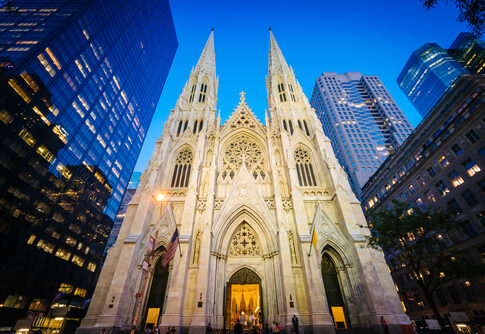 Gothic cathedral illuminated at dusk surrounded by modern skyscrapers